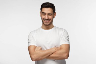 Gülümseyen ve göz kırpıyor, çapraz kollu yakışıklı adam gri arka plan üzerinde izole kamera bakıyor