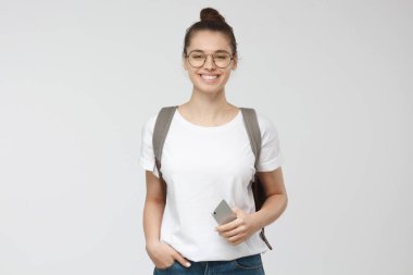 Genç kadın eller cepler poz ayakta gülümseyen, gri arka plan üzerinde izole beyaz tshirt ve trasparent gözlük