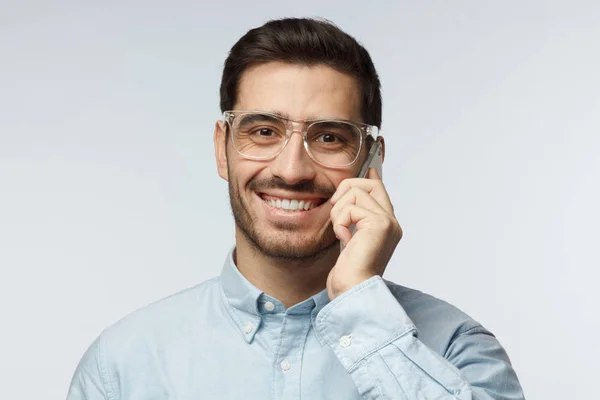 Vértes lövés a fiatal férfi élvezi telefonbeszélgetés toothy boldog mosollyal, nevetve elegáns átlátszó szemüveget visel, szürke háttér elszigetelt — Stock Fotó