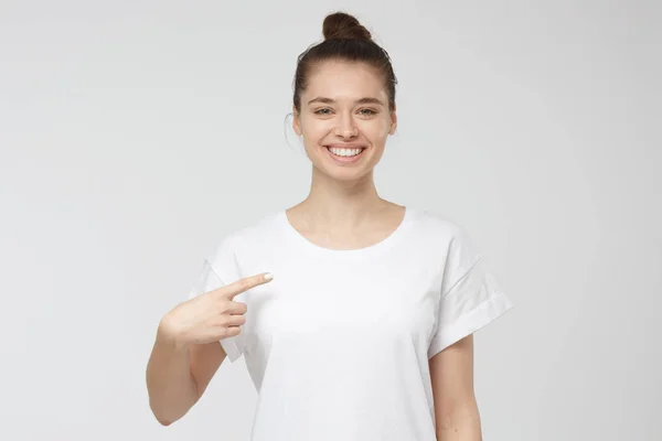 Fiatal nő mutatva a mutatóujját az üres fehér póló, üres hely a hirdetési szöveget vagy képet, állandó elszigetelt szürke háttér — Stock Fotó