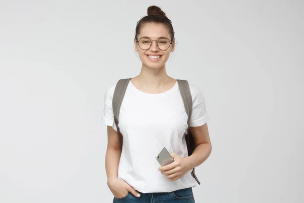 年轻女子在白色的 t恤和 trasparent 眼镜孤立在灰色的背景下, 微笑着, 站在手中的口袋姿势 — 图库照片