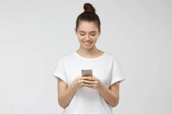 Binnen schot van knappe jonge vrouw die geïsoleerd op grijze achtergrond op smartphone, lacht openlijk smartphone in beide handen houd — Stockfoto