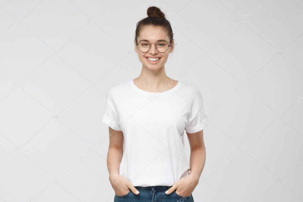 Young european woman standing with hands in pockets, wearing blank white tshirt with copy space for your logo or text, isolated on grey background