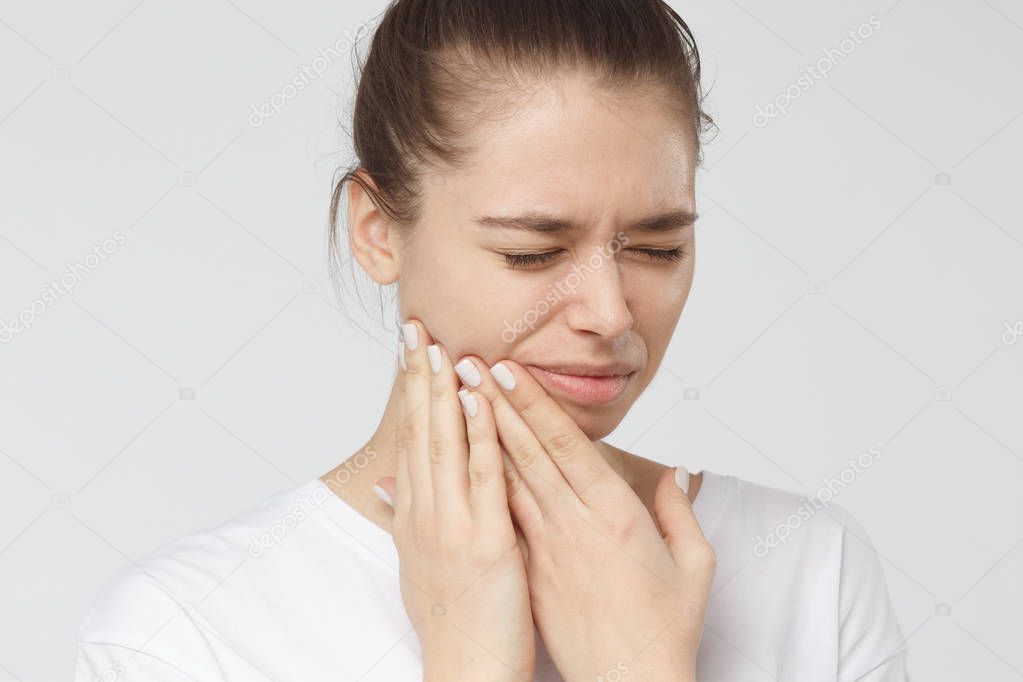 Closeup of beautiful young woman isolated on grey background touching her face and closing eyes with expression of horrible suffer from health problem and aching tooth, showing dissatisfaction.