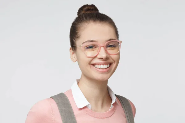 Retrato interior de una hermosa estudiante universitaria con gafas rosas de moda y ropa clásica neutra con cabello castaño atado en moño, sonriente y dispuesta a expresar opiniones — Foto de Stock