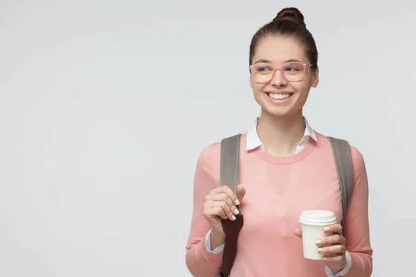 美丽的年轻女性的室内照片, 在粉红色的眼镜上孤立的灰色背景与袋子在她的背上, 微笑着, 拿着一杯咖啡去。Copyspace — 图库照片