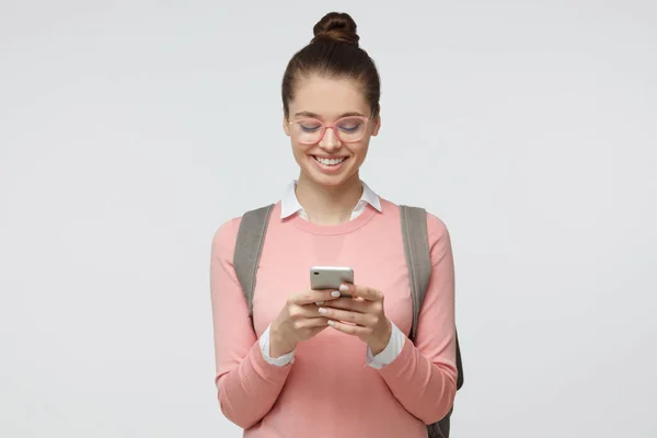 Indoor tiro de boa aparência estudante do sexo feminino isolado em fundo cinza olhando para o smartphone, sorrindo abertamente enquanto segurando smartphone em ambas as mãos — Fotografia de Stock