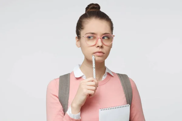 Nahaufnahme Porträt einer jungen Frau, die isoliert auf grauem Hintergrund steht, eine Brille trägt, den Bleistift neben die Lippen hält, nach links denkt und sucht, um Antwort oder Lösung zu finden. — Stockfoto