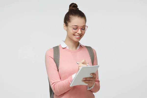 Innenaufnahme einer jungen Frau mit Brille vor grauem Hintergrund, die Notizen auf leere Blätter des Schreibblocks macht, die sie mit zufriedenem und interessiertem Lächeln in der Hand hält, glücklich, zu studieren — Stockfoto