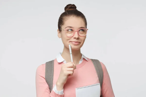 Menina estudante bonita em fundo cinza está pronto para escrever novas informações com caneta, cabelo amarrado em coque, olhando sonhador e entusiasmado com a aprendizagem — Fotografia de Stock