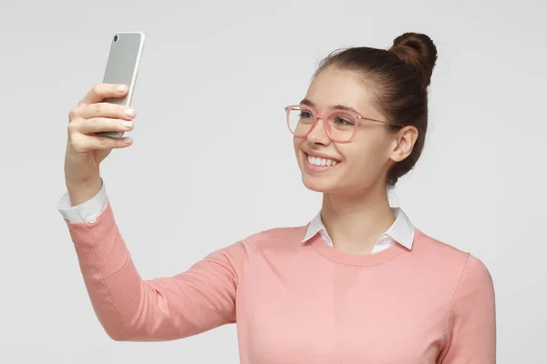 Retrato de jovem mulher em pé contra fundo cinza usando smartphone para tirar fotos de selfie e sorrir — Fotografia de Stock