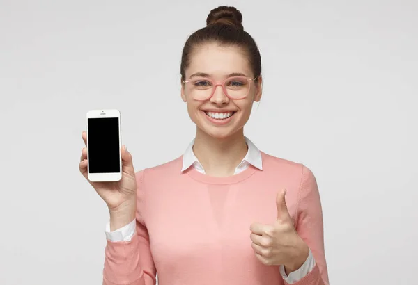 Retrato interno de menina atraente isolado em fundo cinza, segurando smartphone em branco, sorrindo para a câmera, mostrando thubms up gesto, sentindo-se feliz — Fotografia de Stock