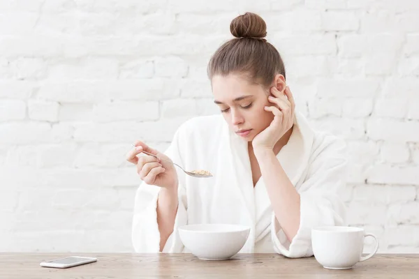 Retrato de jovem mulher entediada comendo cereais com mau gosto em sua cozinha loft branco, sentado à mesa de madeira. Conceito aborrecido da vida cotidiana — Fotografia de Stock
