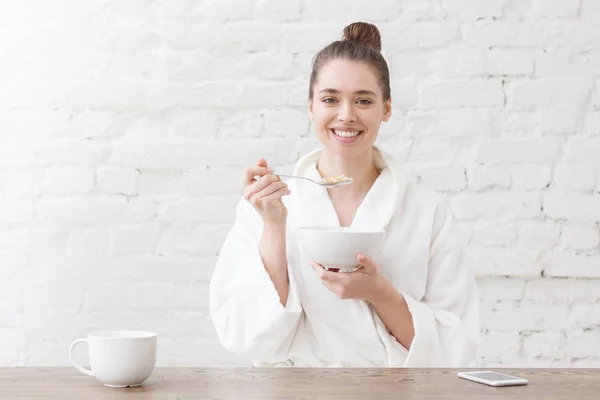 Muito sorridente jovem mulher olhando para a câmera e tomando café da manhã, segurando tigela com cereais com a mão, enquanto sentado à mesa de madeira na cozinha loft branco — Fotografia de Stock