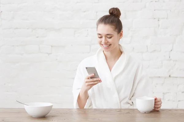 Jovem mulher sentada na cozinha loft branco, após o banho, olhando para o seu smartphone, tendo delicioso café da manhã, verificando mensagens, segurando xícara de café em uma mão — Fotografia de Stock