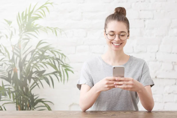 Jovem empresária sorridente em camiseta cinza trabalhando online com smartphone enquanto se senta na mesa de madeira em seu escritório em casa — Fotografia de Stock