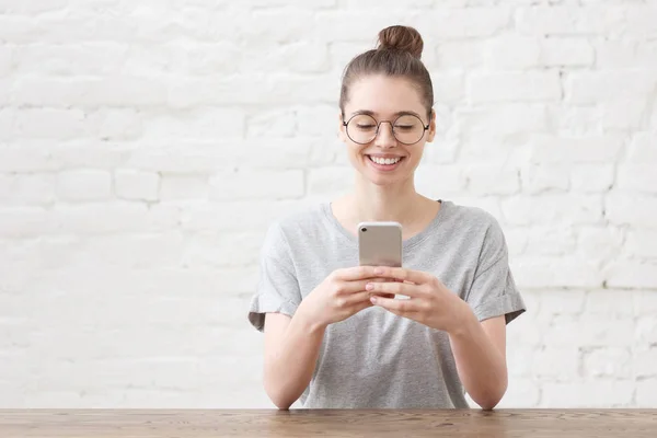 Mujer creativa lleva elegantes gafas redondas y camiseta gris, se sienta en la oficina, utiliza un teléfono inteligente moderno, sonríe felizmente, aislado sobre la pared de ladrillo blanco. Copiar espacio en el lado izquierdo —  Fotos de Stock