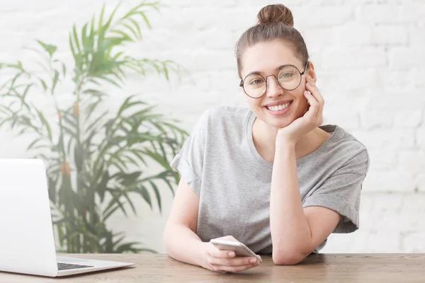 Young hipster woman with bun checking her news feed or messaging, using free wi-fi on her smartphone, smiling at camera — Stock Photo, Image
