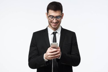 Akıllı telefon ekranına bakarak, finans uygulamasını kullanarak ve gülümseyen güzel gri arka plan üzerinde duran genç iş adamı izole 