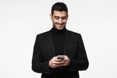 Cep telefonu ekranına dikkatle bakarak, web sayfalarını tarama ve güzel sohbet ederken gülümseyen gri arka plan üzerinde duran genç iş adamı izole