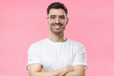 Yakışıklı adam beyaz t-shirt ve çapraz kolları üzerinde pembe arka plan izole olan ayakta şeffaf gözlük gülümseyen portresi