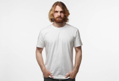 Elleri ceplerinde duran, logo veya metin, gri arka plan üzerinde izole kopya alanı olan beyaz tshirt giyen genç Avrupa erkeksi adam