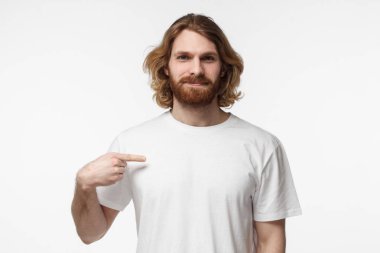 Genç adam gri arka plan üzerinde izole reklam metni veya resmi, ayakta için boş alan ile boş beyaz tshirt, işaret parmağı ile işaret eden sakallı