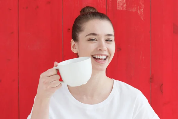 Retrato de jovem mulher em tshirt branca com grande xícara de café na mão, sorrindo amplamente, de pé contra o fundo vermelho — Fotografia de Stock