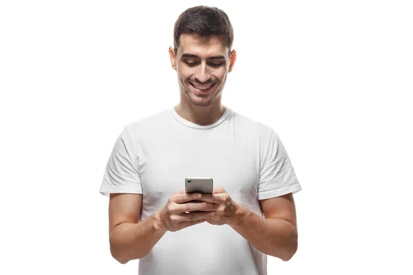 Молодой человек в пустой футболке стоял изолированный на белом фоне, внимательно глядя на экран мобильного телефона, просматривая веб-страницы и красиво улыбаясь во время общения с друзьями — стоковое фото