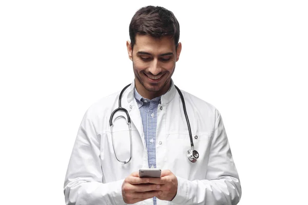 Foto de primer plano del médico hombre de pie aislado sobre fondo blanco mirando atentamente a la pantalla del teléfono celular, sonriendo muy bien —  Fotos de Stock