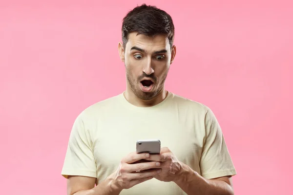 Porträt eines jungen Mannes isoliert auf rosa Hintergrund, der aufgeregt auf das Display seines Smartphones blickt, beeindruckt von Medieninhalten aus dem Web — Stockfoto