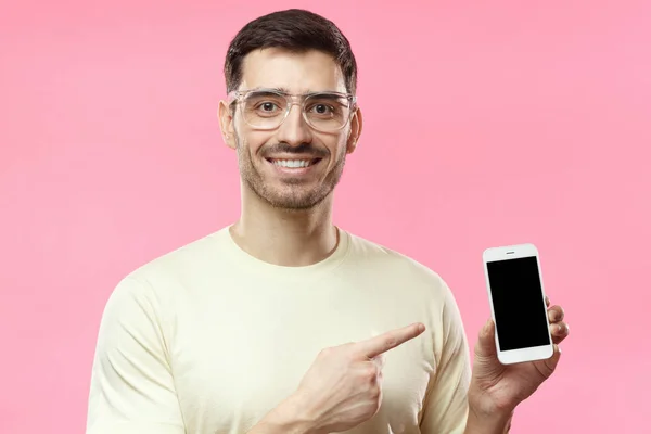 Retrato interno de jovem atraente vestindo óculos transparentes isolados em fundo rosa, segurando smartphone em branco, sorrindo para a câmera, sentindo-se feliz — Fotografia de Stock