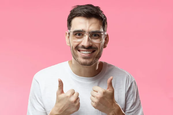 Мотивированный возбужденный молодой человек в белой футболке делает большой палец вверх жест одобрения и успеха с улыбкой, изолированные на розовом фоне — стоковое фото