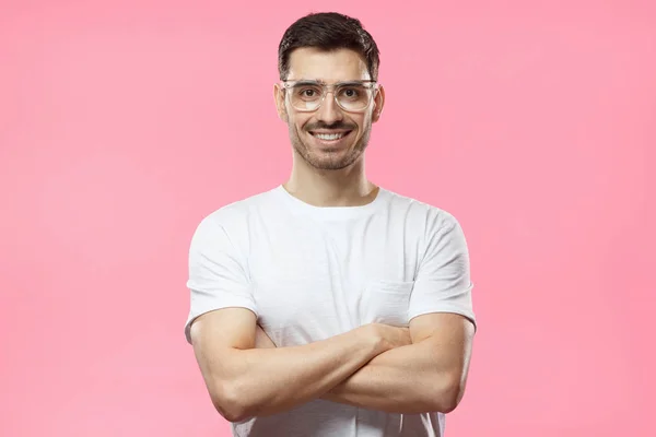 Портрет улыбающегося красивого мужчины в прозрачных очках и белой футболке, стоящей со скрещенными руками на розовом фоне — стоковое фото