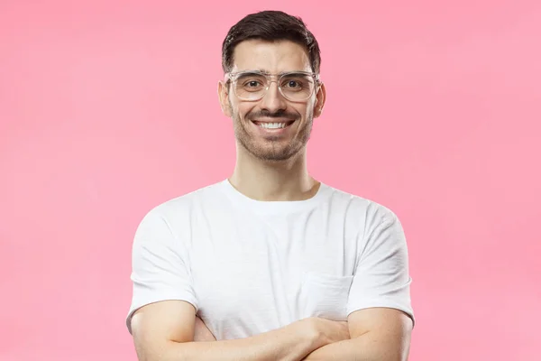 Mosolyogva jóképű férfi fehér póló és állandó keresztezett karokkal elszigetelt rózsaszín háttér átlátszó szemüveget portréja — Stock Fotó