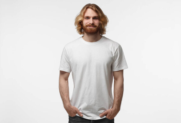 Young european smiling man standing with hands in pockets, wearing blank white tshirt with copy space isolated on grey background