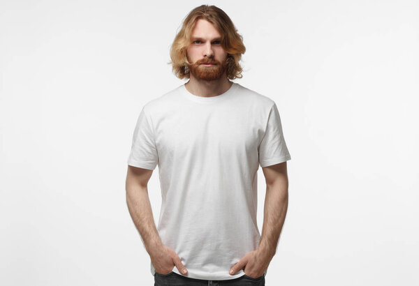 Young european masculine man standing with hands in pockets, wearing white tshirt with copy space for your logo or text, isolated on grey background
