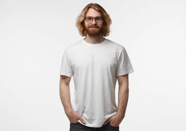 Handsome man in white tshirt and eyeglasses isolated on grey background, smiling, standing with hands in pockets — Stock Photo, Image