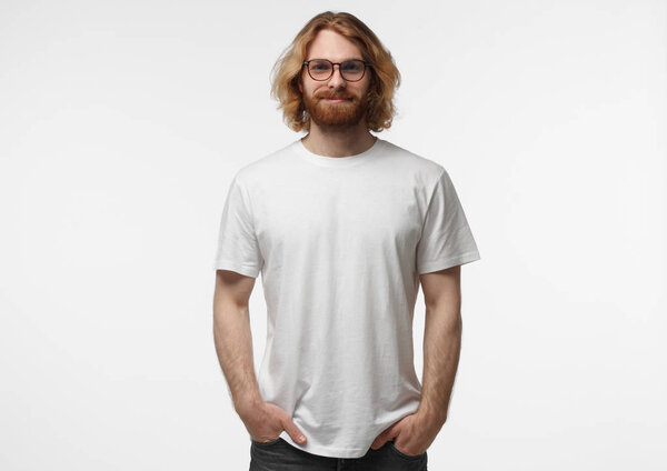 Handsome man in white tshirt and eyeglasses isolated on grey background, smiling, standing with hands in pockets