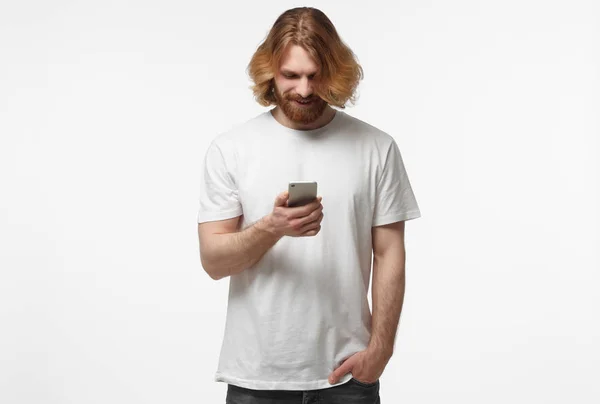 Young man in white tshirt standing isolated on gray background looking attentively at screen of cellphone, browsing web pages and smiling nicely while chatting with friends — Stock Photo, Image
