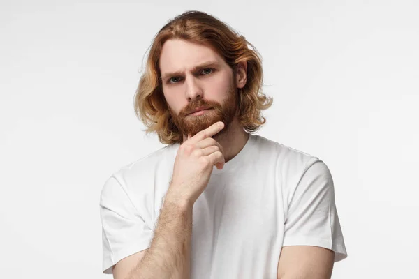 Close up portrait of young handsome bearded man in white tshirt touching his chin with hand thoughtfully looking at camera. Doubt concept — Stock Photo, Image