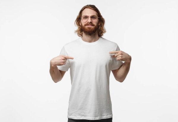 Bearded young man in trendy glasses pointing to his blank white tshirt with index fingers, showing empty space for your advertising text or logo, standing isolated on grey background