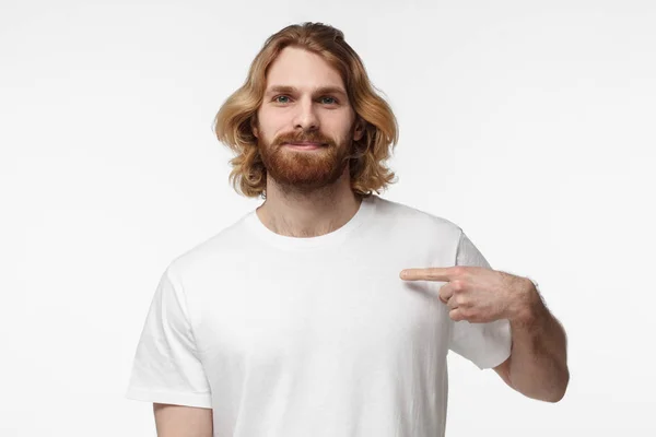 Smiling nice bearded man pointing at his blank white tshirt with index finger, copy space for your advertising, isolated on grey background — Stock Photo, Image