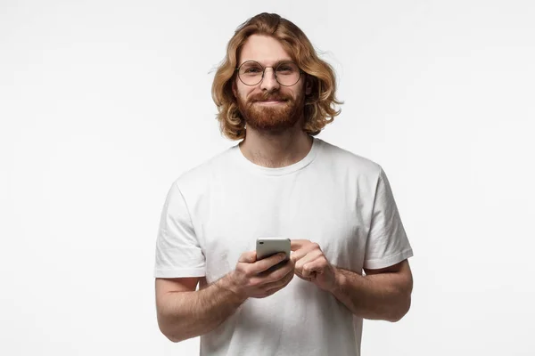 Portret przystojny młody Brodaty mężczyzna w biały tshirt i okrągłe okulary, trzymając smartfon, patrząc na kamery i uśmiecha się na białym tle na szarym tle — Zdjęcie stockowe