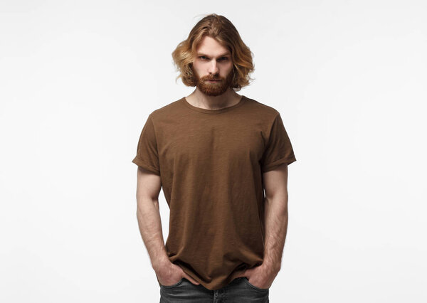 Young european masculine bearded man standing with hands in pockets, in brown tshirt, isolated on grey background