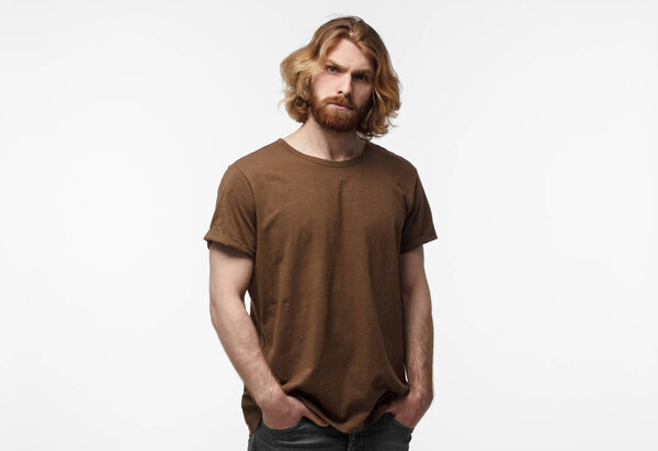 Young brutal bearded man with long hair standing with hands in pockets, in brown tshirt, isolated on grey background