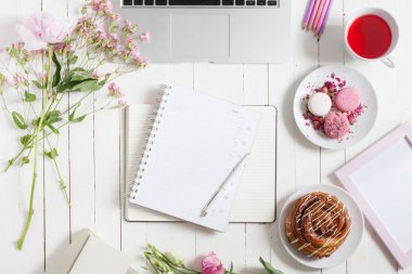 Düz üst çalışma tablosunun görünümünü pembe çiçekler, macarons, çay, bun ve renkli kalemler ile metal dizüstü bilgisayar ile beyaz boyalı ve kalemle notlar ve planları almak için hazır boş copybooks merkezinde açık