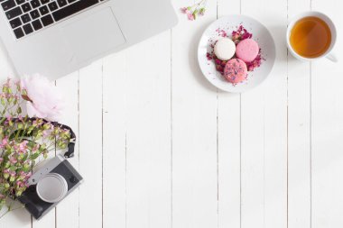 Beyaz ahşap masa gri açık metal dizüstü bilgisayar, pembe çiçekler, retro ince tabaka fotoğraf makinesi, çay bardağı ve tabağı ile boşaltmak reklam yerleştirmek için ortada pembe macarons ile izole düz görünüm