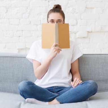 Genç Avrupa bayanın ilginç bir şeyler saklamaya kitap kapağı ile yüzün alt kısmını gizleme üzerinde gözlüklü beyaz tuğla arka plan gri Divan resimde kare resim o okumuş