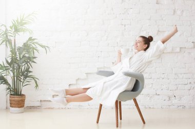 Studio photo of young good-looking European Caucasian female pictured in bright white room dressed in white bathrobe stretching having just woken and got up in morning getting ready for new day clipart